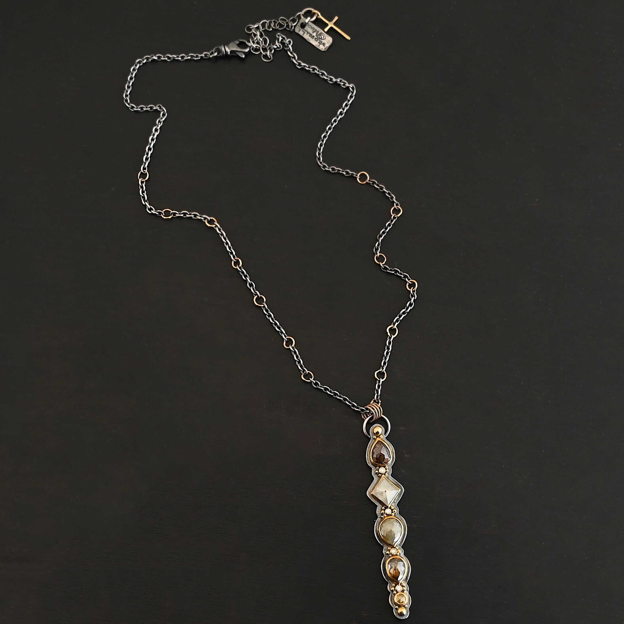 Daybreak Necklace Photo of the "Daybreak" Necklace. Made out of sterling silver, gold, and diamond. There are nine diamonds, all in a straight line, that create the pendant of the necklace. Five of the diamonds are different shapes, sizes, and colors. Each of these diamonds are separated by small, circular diamonds, which there are four of. The top of the pendant starts with a large diamond, then a small one, and keeps alternating until the final diamond is a large one. There is a large loop soldered to the back of the top of the pendant with four gold jump rings attached to it. Going through the jump rings is the silver chain of the necklace. The chain of the necklace has gold loops scattered around it. Necklace is lying against a dark background.