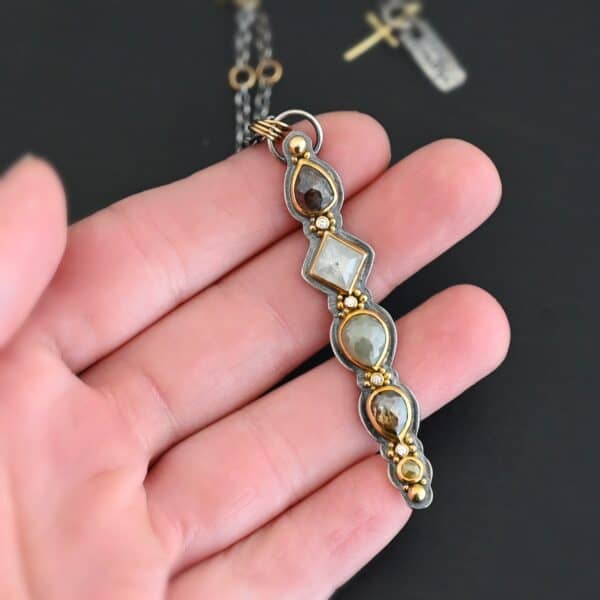 Photo of the "Daybreak" Necklace. Made out of sterling silver, gold, and diamond. There are nine diamonds, all in a straight line, that create the pendant of the necklace. Five of the diamonds are different shapes, sizes, and colors. Each of these diamonds are separated by small, circular diamonds, which there are four of. The top of the pendant starts with a large diamond, then a small one, and keeps alternating until the final diamond is a large one. There is a large loop soldered to the back of the top of the pendant with four gold jump rings attached to it. Going through the jump rings is the silver chain of the necklace. The chain of the necklace has gold loops scattered around it. Necklace is lying against the fingertips of a hand.