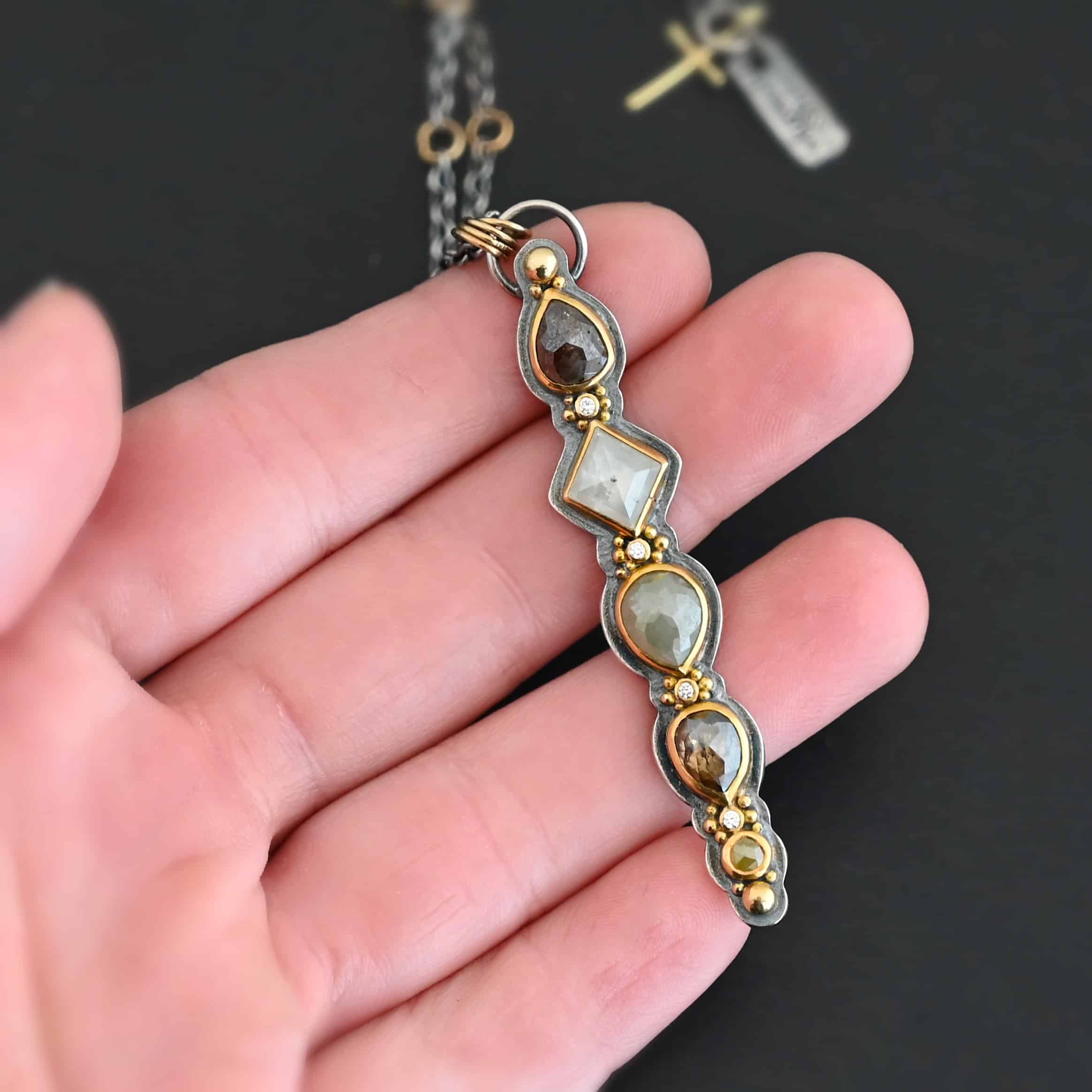 Daybreak Necklace Photo of the "Daybreak" Necklace. Made out of sterling silver, gold, and diamond. There are nine diamonds, all in a straight line, that create the pendant of the necklace. Five of the diamonds are different shapes, sizes, and colors. Each of these diamonds are separated by small, circular diamonds, which there are four of. The top of the pendant starts with a large diamond, then a small one, and keeps alternating until the final diamond is a large one. There is a large loop soldered to the back of the top of the pendant with four gold jump rings attached to it. Going through the jump rings is the silver chain of the necklace. The chain of the necklace has gold loops scattered around it. Necklace is lying against the fingertips of a hand.