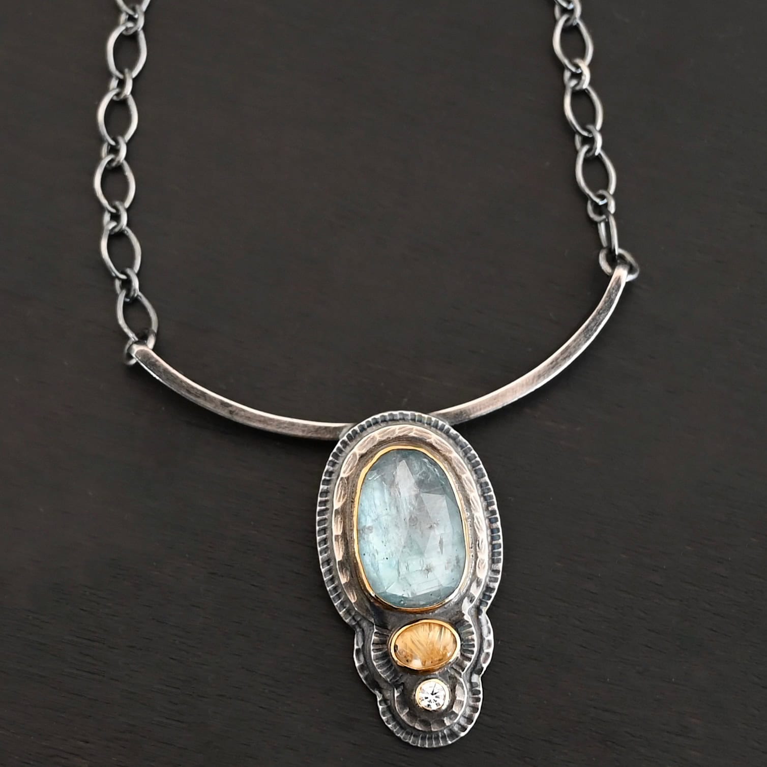 Chasing Waterfalls Necklace Photo of the "Chasing Waterfalls" Necklace. Made out of sterling silver, aquamarine, rutilated quartz, and diamond. The aquamarine is a large oval placed vertically on the top. Beneath this is a small oval rutilated quartz plated horizontally. Finally, there is a small circular diamond beneath the rutilated quartz. Each stone is set in gold. There are two layers of sterling silver. Both layers are hammered, but with different patterns. Pendant is soldered to a curved bar that is smooth and flat. Each end of the curved bar has a loop on it. Attached to each loop on the end is the chain of the necklace. Necklace is lying against a dark background.
