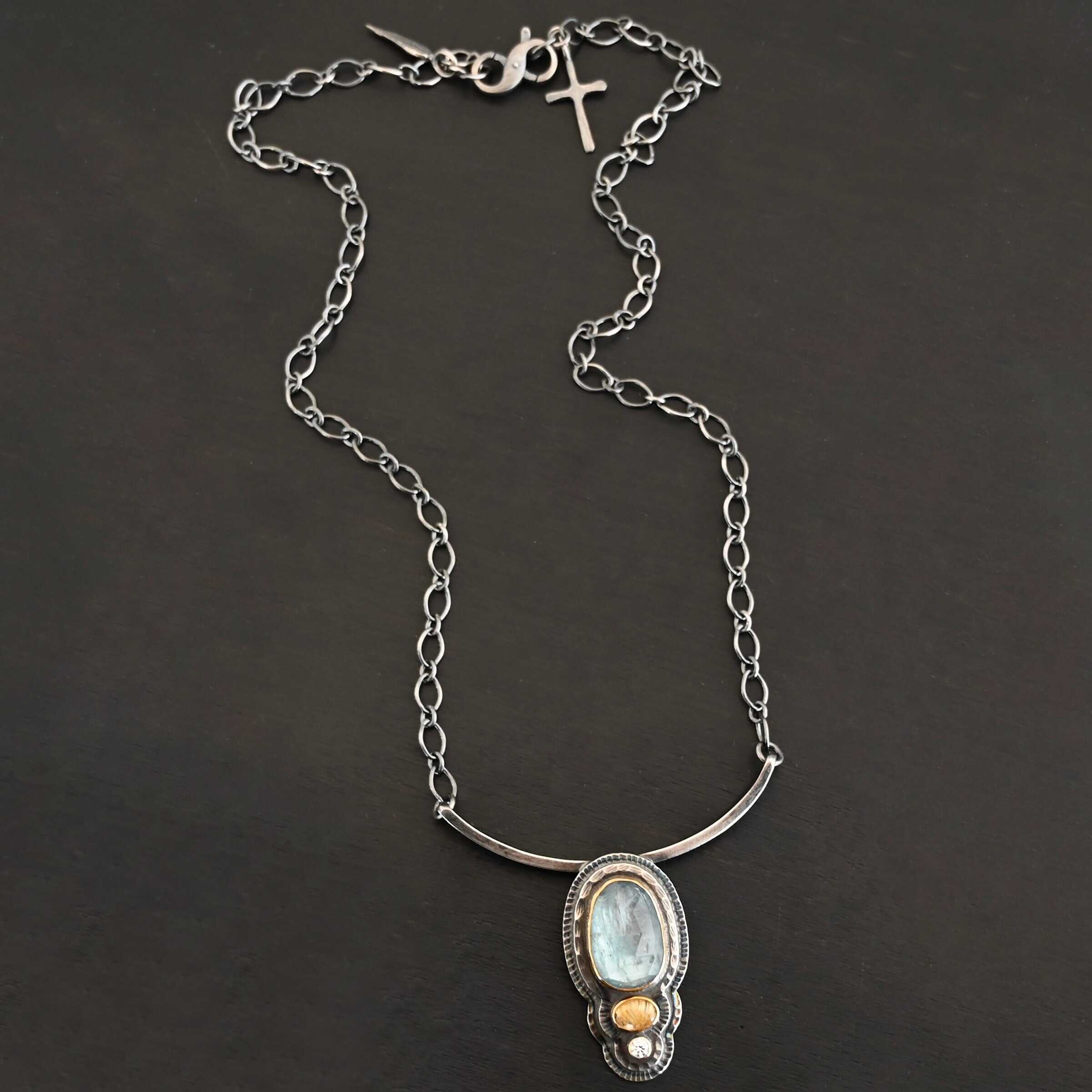 Chasing Waterfalls Necklace Photo of the "Chasing Waterfalls" Necklace. Made out of sterling silver, aquamarine, rutilated quartz, and diamond. The aquamarine is a large oval placed vertically on the top. Beneath this is a small oval rutilated quartz plated horizontally. Finally, there is a small circular diamond beneath the rutilated quartz. Each stone is set in gold. There are two layers of sterling silver. Both layers are hammered, but with different patterns. Pendant is soldered to a curved bar that is smooth and flat. Each end of the curved bar has a loop on it. Attached to each loop on the end is the chain of the necklace. Necklace is lying against a dark background.