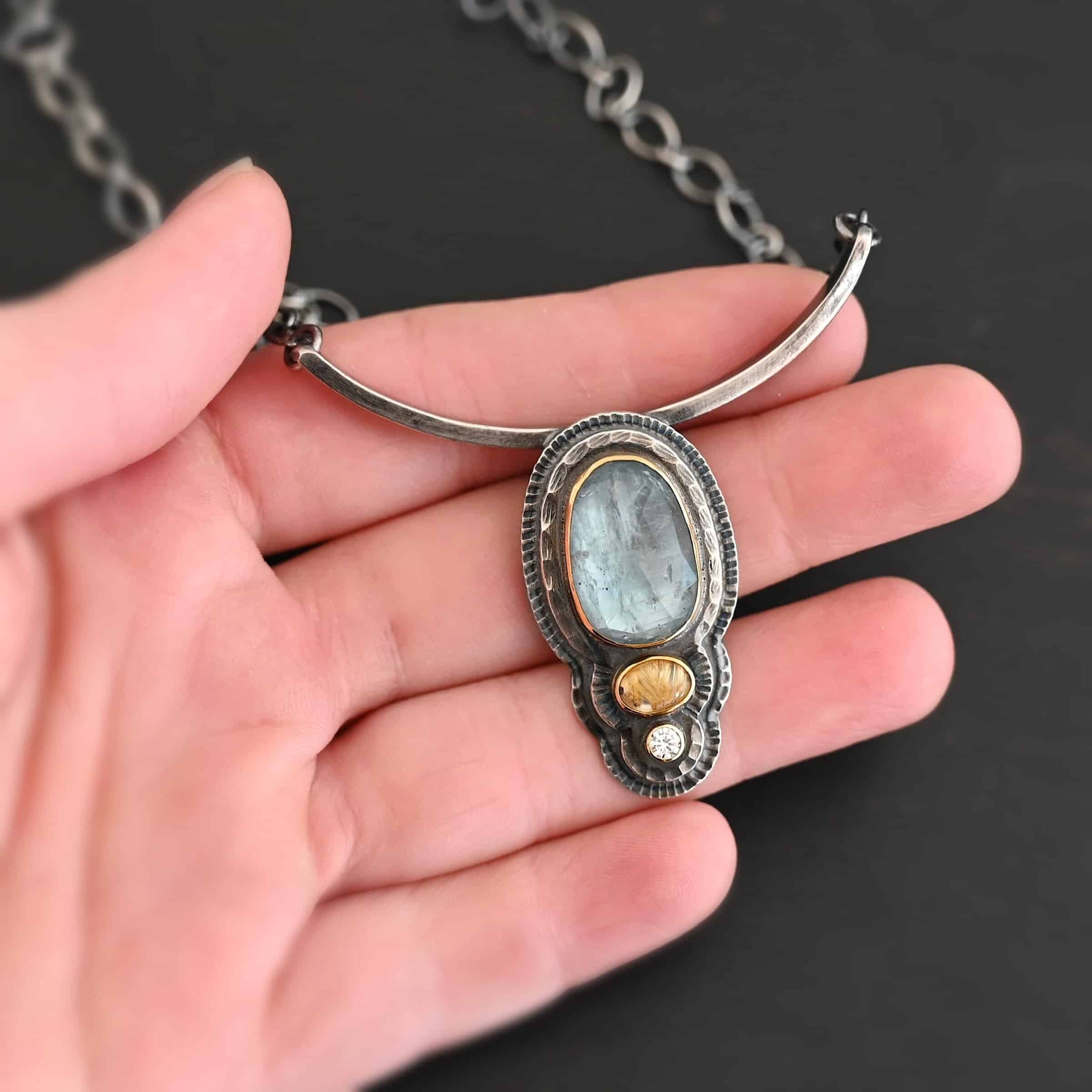 Chasing Waterfalls Necklace Photo of the "Chasing Waterfalls" Necklace. Made out of sterling silver, aquamarine, rutilated quartz, and diamond. The aquamarine is a large oval placed vertically on the top. Beneath this is a small oval rutilated quartz plated horizontally. Finally, there is a small circular diamond beneath the rutilated quartz. Each stone is set in gold. There are two layers of sterling silver. Both layers are hammered, but with different patterns. Pendant is soldered to a curved bar that is smooth and flat. Each end of the curved bar has a loop on it. Attached to each loop on the end is the chain of the necklace. Necklace is lying against the fingertips of a hand.