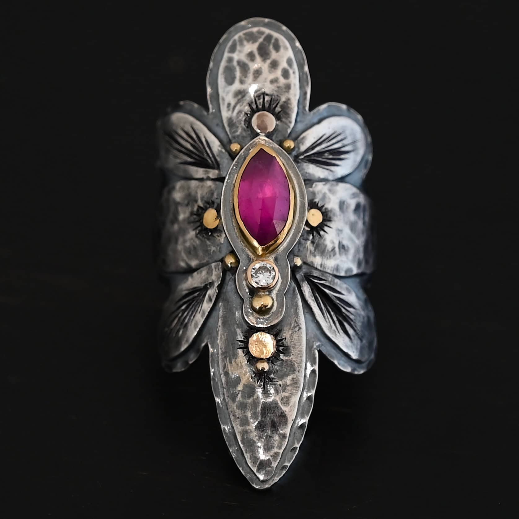 Heaven In A Wildflower Ring Photo of the "Heaven In A Wildflower" Ring. Made out of sterling silver, gold, ruby, and diamond. There is a marquise shaped ruby in the center of the ring with a small circle diamond beneath it. There is a gold ball on the bottom of the diamond and a hammered silver dot on the top of the marquise. The marquise is placed vertically. There is gold ball detail around the rest of the ring, as well as hammered gold dots. The ring is large and long. The entire ring is shaped like multiple flower petals. There is a large flower petal on top that is rounded, six underneath that flower petal (three on each side), and one on the bottom that is pointed. There are two layers of silver. They are both hammered, but with different patterns. The top leaves on each side and bottom leaves on each side in between the top and bottom leaves have engraved lines in them. Ring is sitting against a dark background.