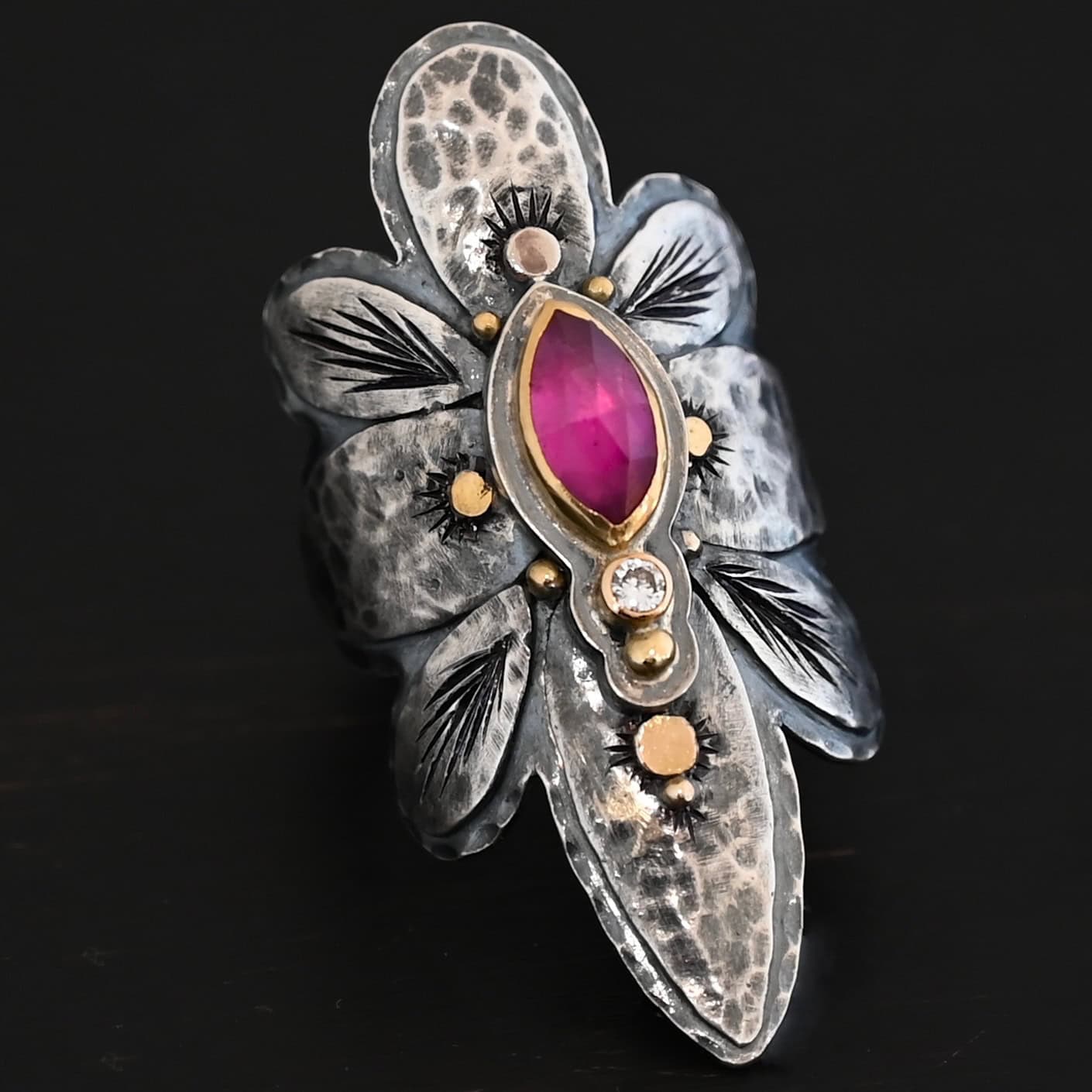 Heaven In A Wildflower Ring Photo of the "Heaven In A Wildflower" Ring. Made out of sterling silver, gold, ruby, and diamond. There is a marquise shaped ruby in the center of the ring with a small circle diamond beneath it. There is a gold ball on the bottom of the diamond and a hammered silver dot on the top of the marquise. The marquise is placed vertically. There is gold ball detail around the rest of the ring, as well as hammered gold dots. The ring is large and long. The entire ring is shaped like multiple flower petals. There is a large flower petal on top that is rounded, six underneath that flower petal (three on each side), and one on the bottom that is pointed. There are two layers of silver. They are both hammered, but with different patterns. The top leaves on each side and bottom leaves on each side in between the top and bottom leaves have engraved lines in them. Ring is sitting against a dark background.