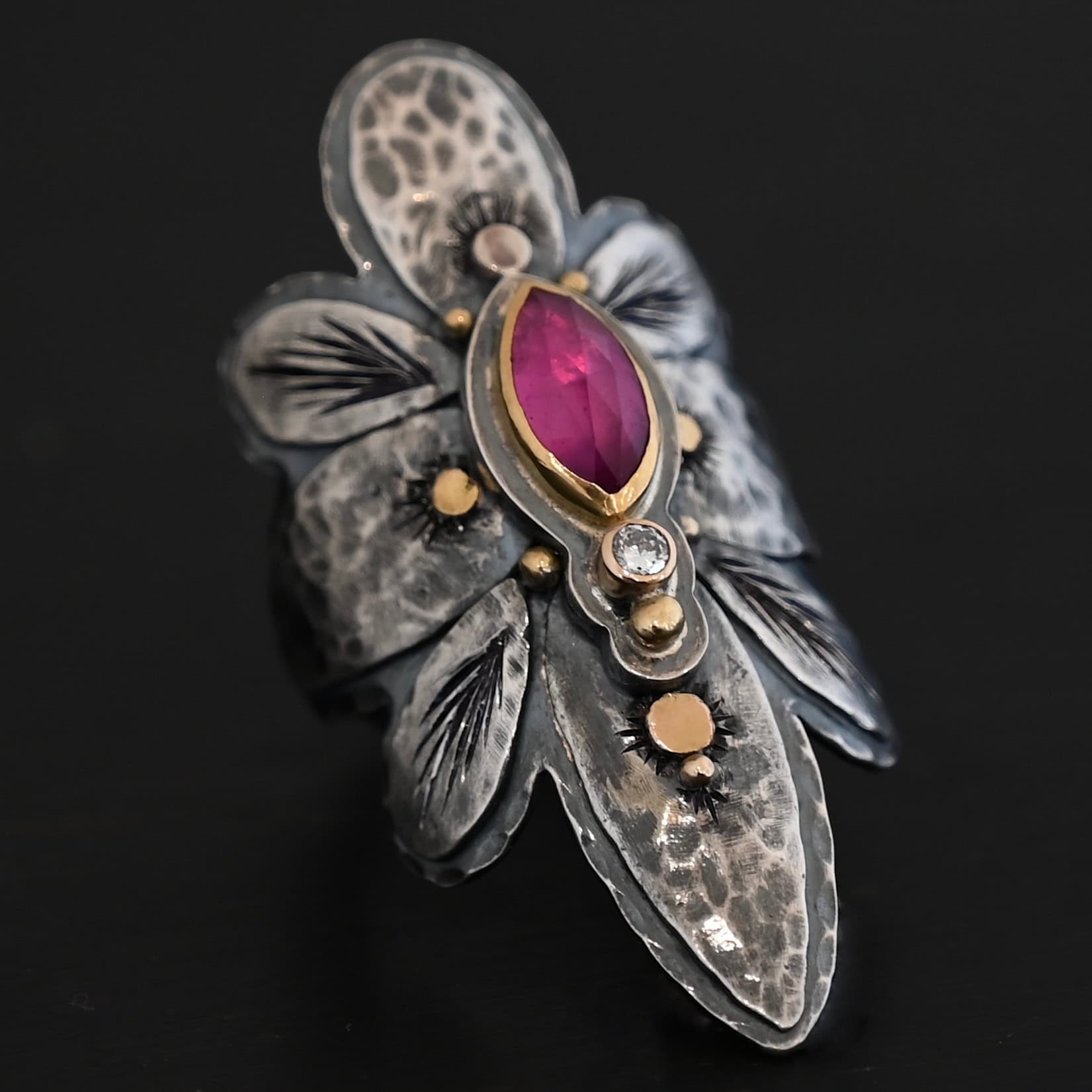 Heaven In A Wildflower Ring Photo of the "Heaven In A Wildflower" Ring. Made out of sterling silver, gold, ruby, and diamond. There is a marquise shaped ruby in the center of the ring with a small circle diamond beneath it. There is a gold ball on the bottom of the diamond and a hammered silver dot on the top of the marquise. The marquise is placed vertically. There is gold ball detail around the rest of the ring, as well as hammered gold dots. The ring is large and long. The entire ring is shaped like multiple flower petals. There is a large flower petal on top that is rounded, six underneath that flower petal (three on each side), and one on the bottom that is pointed. There are two layers of silver. They are both hammered, but with different patterns. The top leaves on each side and bottom leaves on each side in between the top and bottom leaves have engraved lines in them. Ring is sitting against a dark background.