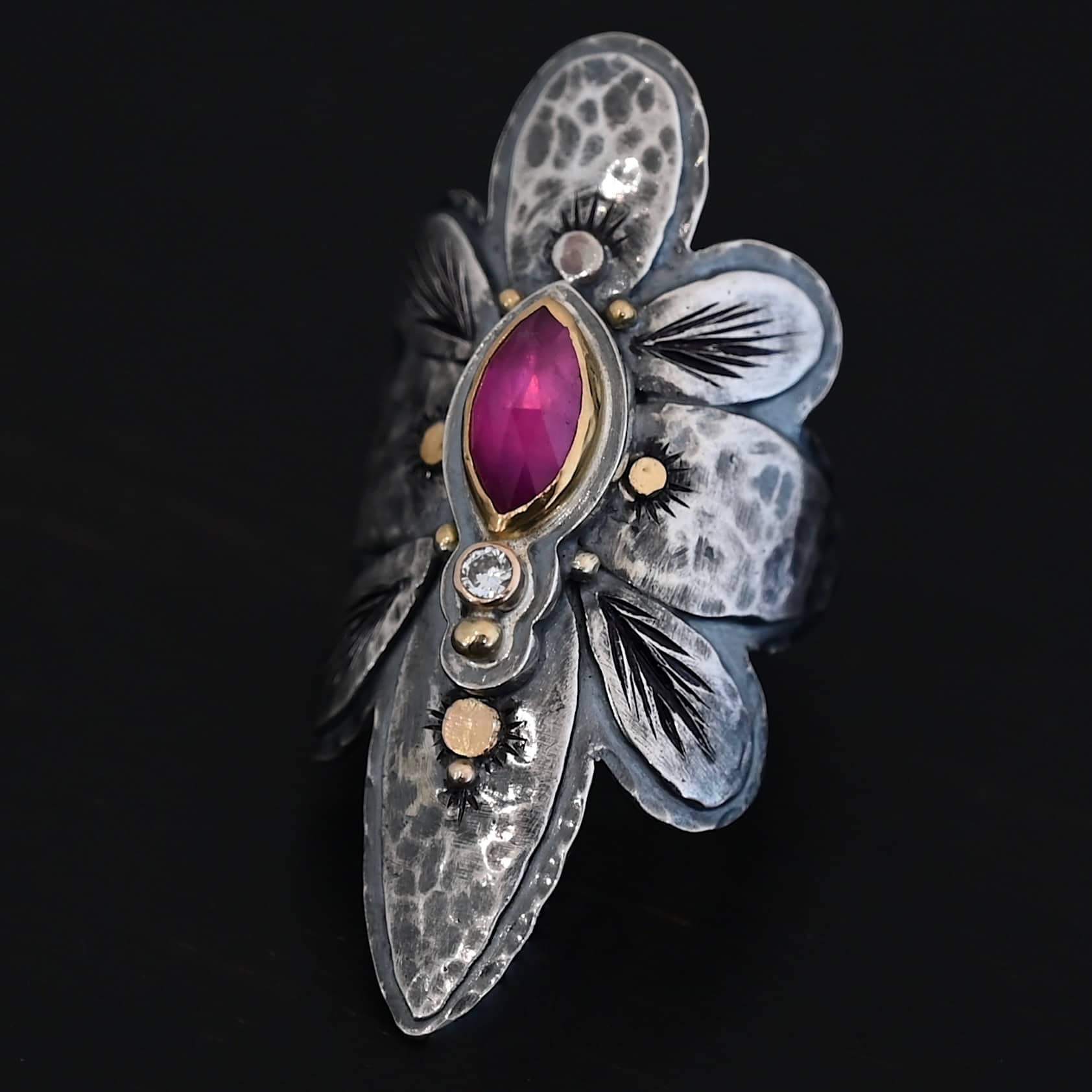Heaven In A Wildflower Ring Photo of the "Heaven In A Wildflower" Ring. Made out of sterling silver, gold, ruby, and diamond. There is a marquise shaped ruby in the center of the ring with a small circle diamond beneath it. There is a gold ball on the bottom of the diamond and a hammered silver dot on the top of the marquise. The marquise is placed vertically. There is gold ball detail around the rest of the ring, as well as hammered gold dots. The ring is large and long. The entire ring is shaped like multiple flower petals. There is a large flower petal on top that is rounded, six underneath that flower petal (three on each side), and one on the bottom that is pointed. There are two layers of silver. They are both hammered, but with different patterns. The top leaves on each side and bottom leaves on each side in between the top and bottom leaves have engraved lines in them. Ring is sitting against a dark background.