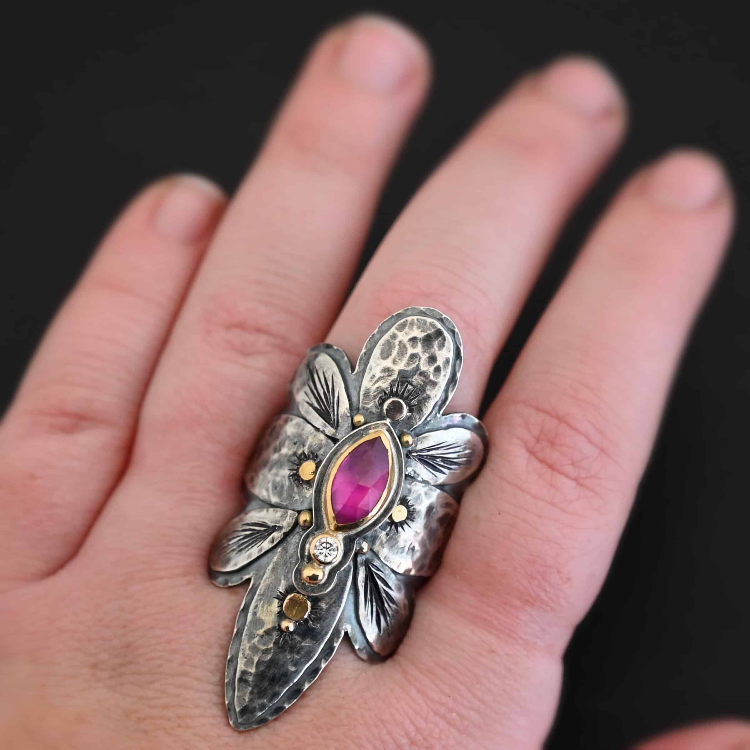 Heaven In A Wildflower Ring Photo of the "Heaven In A Wildflower" Ring. Made out of sterling silver, gold, ruby, and diamond. There is a marquise shaped ruby in the center of the ring with a small circle diamond beneath it. There is a gold ball on the bottom of the diamond and a hammered silver dot on the top of the marquise. The marquise is placed vertically. There is gold ball detail around the rest of the ring, as well as hammered gold dots. The ring is large and long. The entire ring is shaped like multiple flower petals. There is a large flower petal on top that is rounded, six underneath that flower petal (three on each side), and one on the bottom that is pointed. There are two layers of silver. They are both hammered, but with different patterns. The top leaves on each side and bottom leaves on each side in between the top and bottom leaves have engraved lines in them. Ring is worn by a model up close to demonstrate size. Ring is worn on a models middle finger, and goes from the knuckle of the finger past the bottom of the pointer finger to the middle of the thumb.