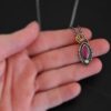 Photo of the "Soft Petals" Necklace. Made out of sterling silver, gold, ruby, and diamond. The bottom of the ring has a small circle diamond. Above this is a pink, marquise shaped ruby placed vertically, so that the points are pointing up and down. The stone is set in gold, and there are two layers of sterling silver beneath it. The top layer of silver goes around the edge of the bottom layer. Both layers are marquise shaped, and the top outside layer is hammered. There is a loop on the back of the necklace in which three gold jump rings go through. Going through the jump rings is the silver chain of the necklace. Necklace is laying against the fingertips of a hand to demonstrate size.