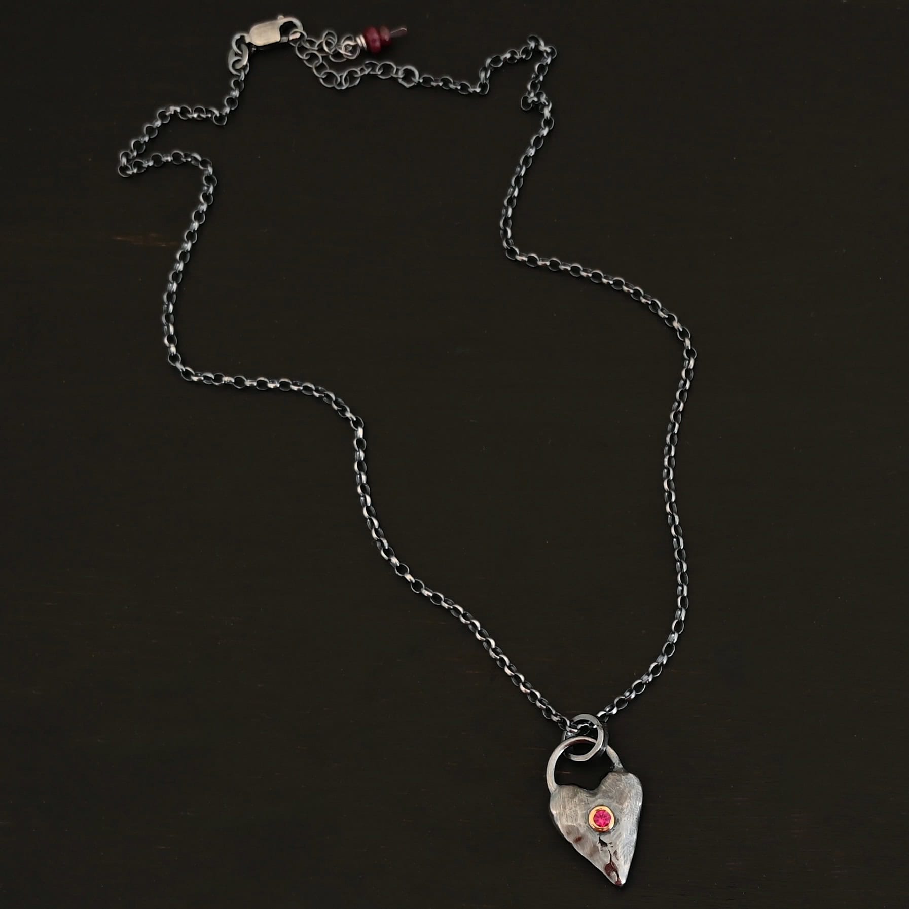 In Your Heart Birthstone Necklace - Ruby Photo of the "In Your Heart" Necklace. Made out of sterling silver and gold with a birthstone. The pendant is a solid sterling silver heart that is hammered and oxidized with a ruby birthstone in the center that is set in gold. There is a silver loop that goes from one side of the heart to the other on the top. Attached to this heart is a large jump ring that is oxidized. Going through this is the silver chain for the necklace. Necklace is laying against a dark background.