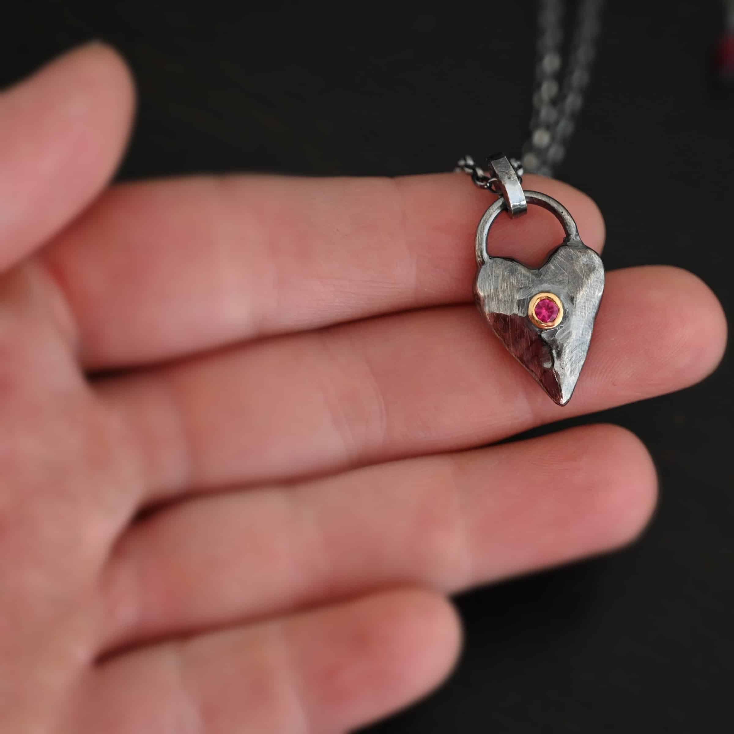 In Your Heart Birthstone Necklace - Ruby Photo of the "In Your Heart" Necklace. Made out of sterling silver and gold with a birthstone. The pendant is a solid sterling silver heart that is hammered and oxidized with a ruby birthstone in the center that is set in gold. There is a silver loop that goes from one side of the heart to the other on the top. Attached to this heart is a large jump ring that is oxidized. Going through this is the silver chain for the necklace. Necklace is laying against the fingertips of a hand to demonstrate size of the pendant.