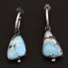 Photo of the "Frozen Land" Earrings shown up close. Earrings are made out of sterling silver and turquoise. Each earring has a triangle shaped turquoise, but the right earring's turquoise is longer than the right earring. Each turquoise is a light blue color with dark brown lines scattered around it. On the bottom of each earring is a silver ball. The top of each earring has a silver loop soldered to it. Going through the loop is the half circle post ear wire. Earrings are laying against a dark background.