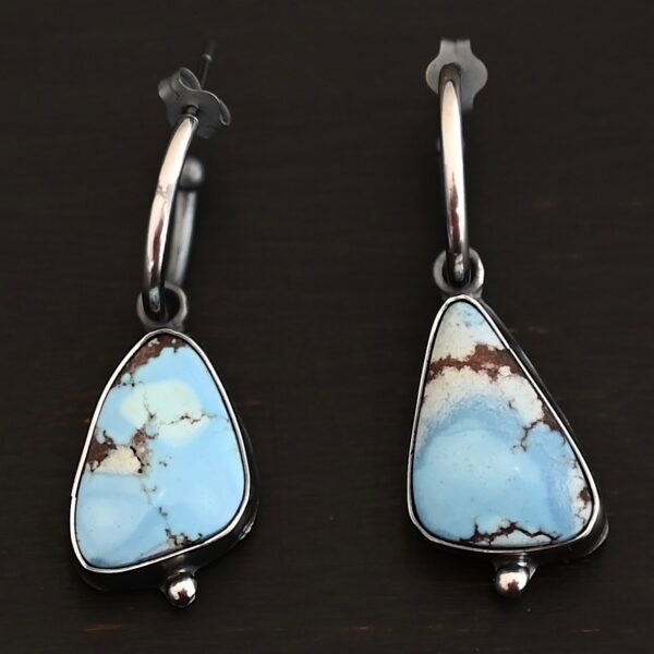 Photo of the "Frozen Land" Earrings shown up close. Earrings are made out of sterling silver and turquoise. Each earring has a triangle shaped turquoise, but the right earring's turquoise is longer than the right earring. Each turquoise is a light blue color with dark brown lines scattered around it. On the bottom of each earring is a silver ball. The top of each earring has a silver loop soldered to it. Going through the loop is the half circle post ear wire. Earrings are laying against a dark background.
