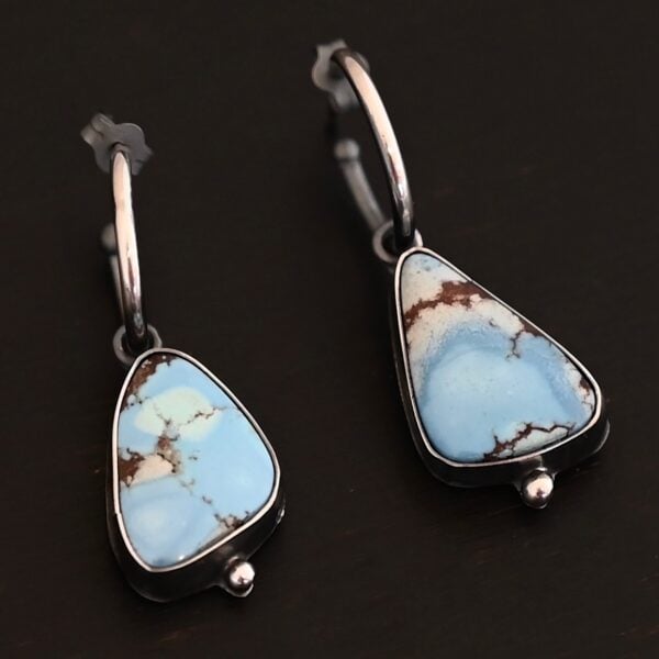 Photo of the "Frozen Land" Earrings shown up close. Earrings are made out of sterling silver and turquoise. Each earring has a triangle shaped turquoise, but the right earring's turquoise is longer than the right earring. Each turquoise is a light blue color with dark brown lines scattered around it. On the bottom of each earring is a silver ball. The top of each earring has a silver loop soldered to it. Going through the loop is the half circle post ear wire. Earrings are laying against a dark background.