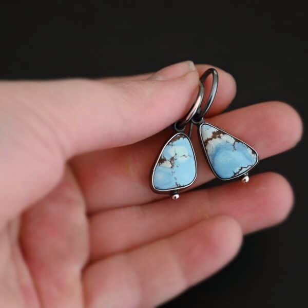 Photo of the "Frozen Land" Earrings shown up close. Earrings are made out of sterling silver and turquoise. Each earring has a triangle shaped turquoise, but the right earring's turquoise is longer than the right earring. Each turquoise is a light blue color with dark brown lines scattered around it. On the bottom of each earring is a silver ball. The top of each earring has a silver loop soldered to it. Going through the loop is the half circle post ear wire. Earrings are held in the fingertips of a hand to demonstrate size.