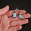 Photo of the "Frozen Land" Earrings shown up close. Earrings are made out of sterling silver and turquoise. Each earring has a triangle shaped turquoise, but the right earring's turquoise is longer than the right earring. Each turquoise is a light blue color with dark brown lines scattered around it. On the bottom of each earring is a silver ball. The top of each earring has a silver loop soldered to it. Going through the loop is the half circle post ear wire. Earrings are laying on the fingertips of a hand to demonstrate size.
