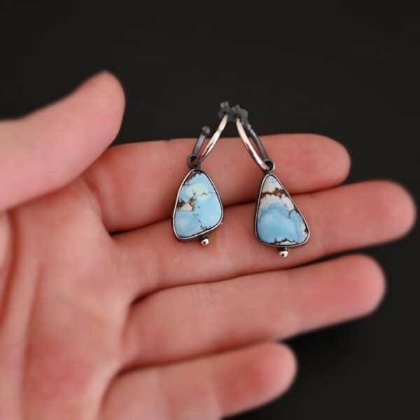 Photo of the "Frozen Land" Earrings shown up close. Earrings are made out of sterling silver and turquoise. Each earring has a triangle shaped turquoise, but the right earring's turquoise is longer than the right earring. Each turquoise is a light blue color with dark brown lines scattered around it. On the bottom of each earring is a silver ball. The top of each earring has a silver loop soldered to it. Going through the loop is the half circle post ear wire. Earrings are laying on the fingertips of a hand to demonstrate size.
