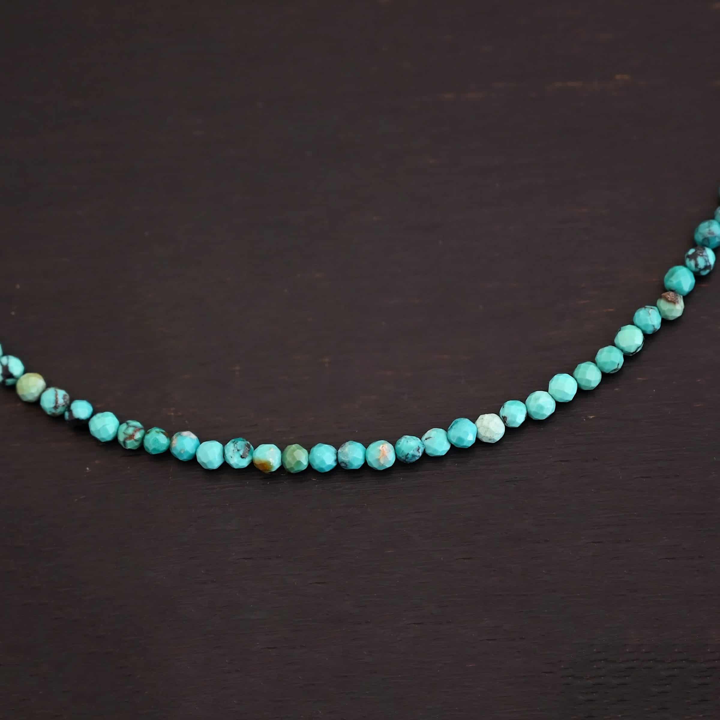 Close up of the "Clara" Necklace. Necklace is made out of smaller sized, spherical, turquoise beads. The beads are strung on heavy wire and are right next to each other tightly. Beads are a mix of blue and green colors. Necklace is lying against a dark background.