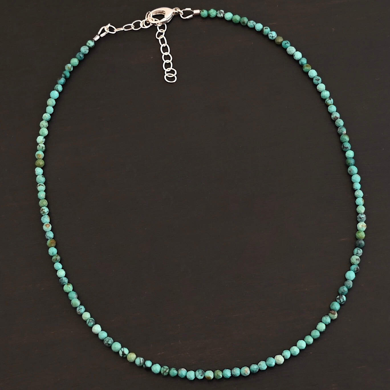 Close up of the "Clara" Necklace. Necklace is made out of smaller sized, spherical, turquoise beads. The beads are strung on heavy wire and are right next to each other tightly. Beads are a mix of blue and green colors. Necklace has a lobster claw clasp and a two inch sterling silver extension. Necklace is lying against a dark background.