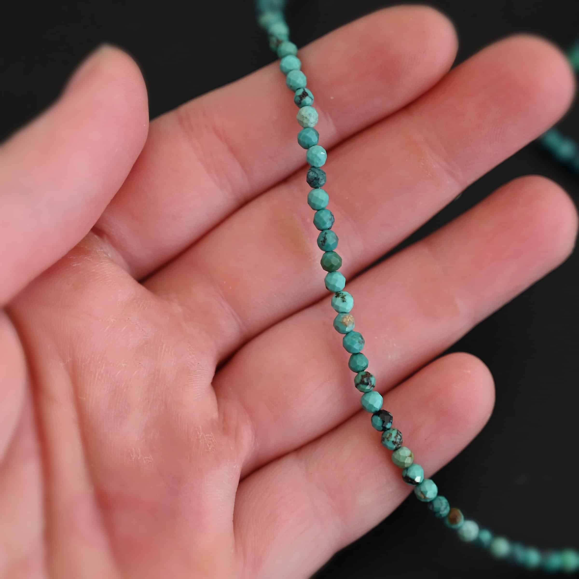 Close up of the "Clara" Necklace. Necklace is made out of smaller sized, spherical, turquoise beads. The beads are strung on heavy wire and are right next to each other tightly. Beads are a mix of blue and green colors. Necklace is lying against the fingertips of a hand to demonstrate size.