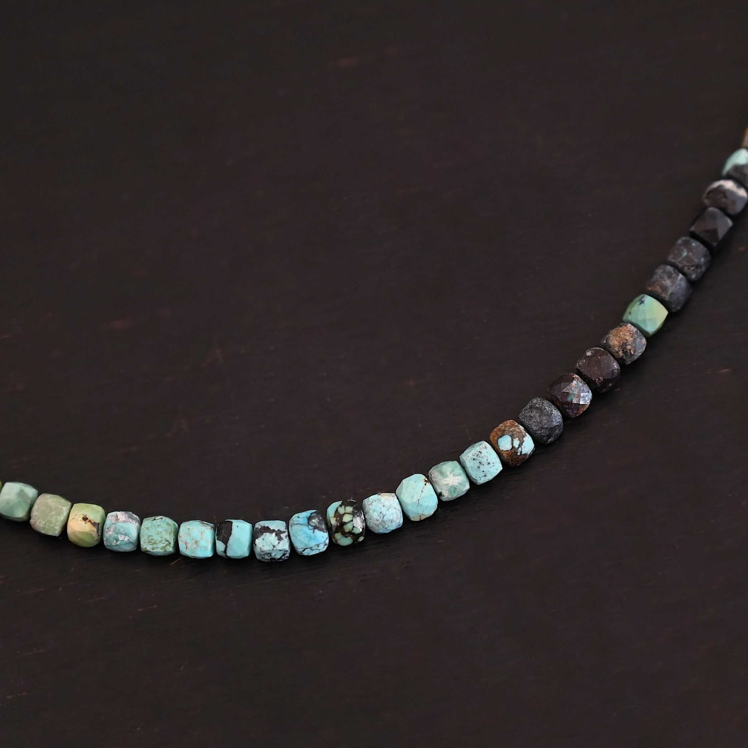 Ada Necklace Close up of the "Ada" Necklace. Necklace is made out of larger sized, square, turquoise beads. The beads are strung on heavy wire and are right next to each other tightly. Beads are a mix of blue, green, black, and brown colors. Necklace is lying against a dark background.