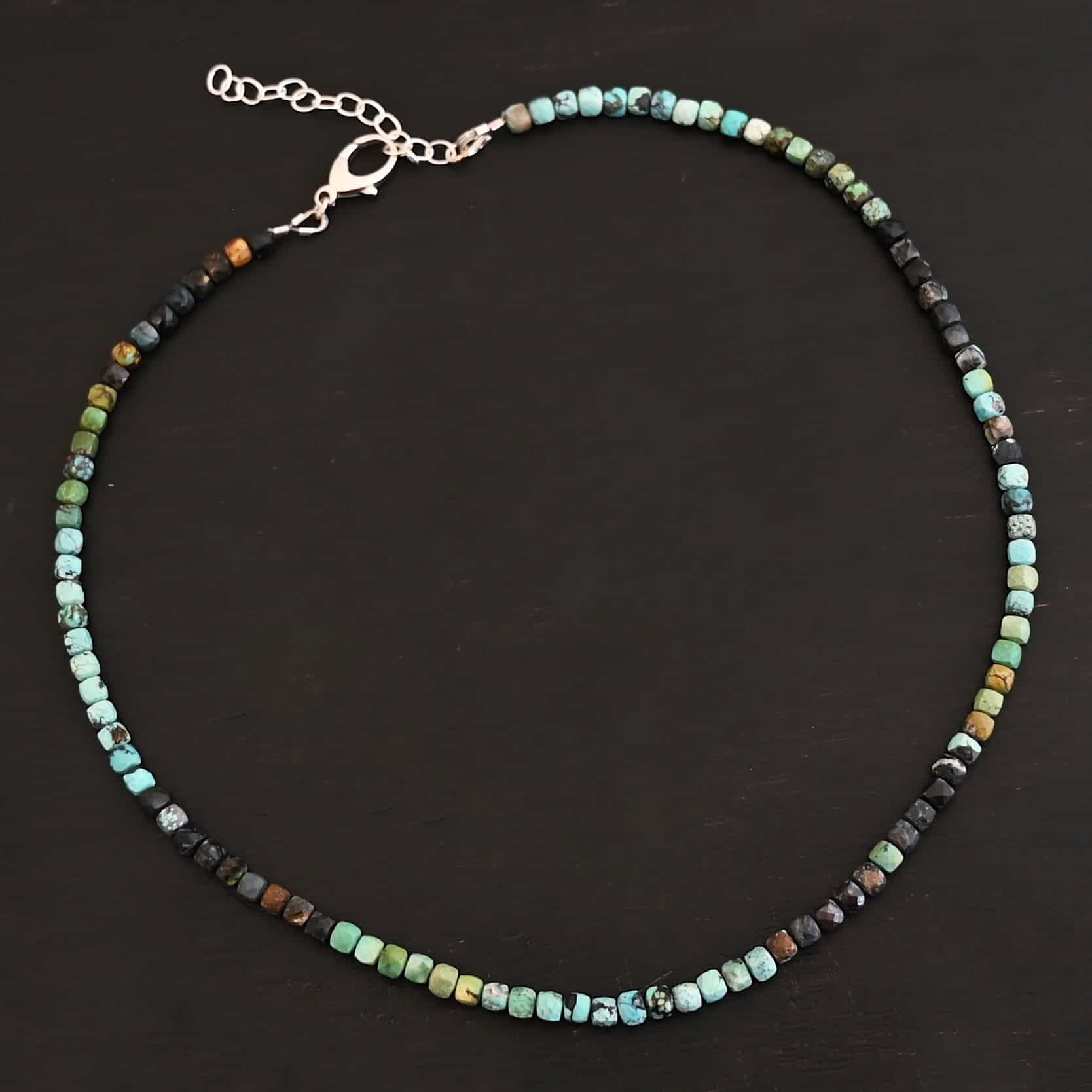 Ada Necklace Close up of the "Ada" Necklace. Necklace is made out of larger sized, square, turquoise beads. The beads are strung on heavy wire and are right next to each other tightly. Beads are a mix of blue, green, black, and brown colors. Necklace has a lobster claw clasp and a two inch sterling silver extension. Necklace is lying against a dark background.