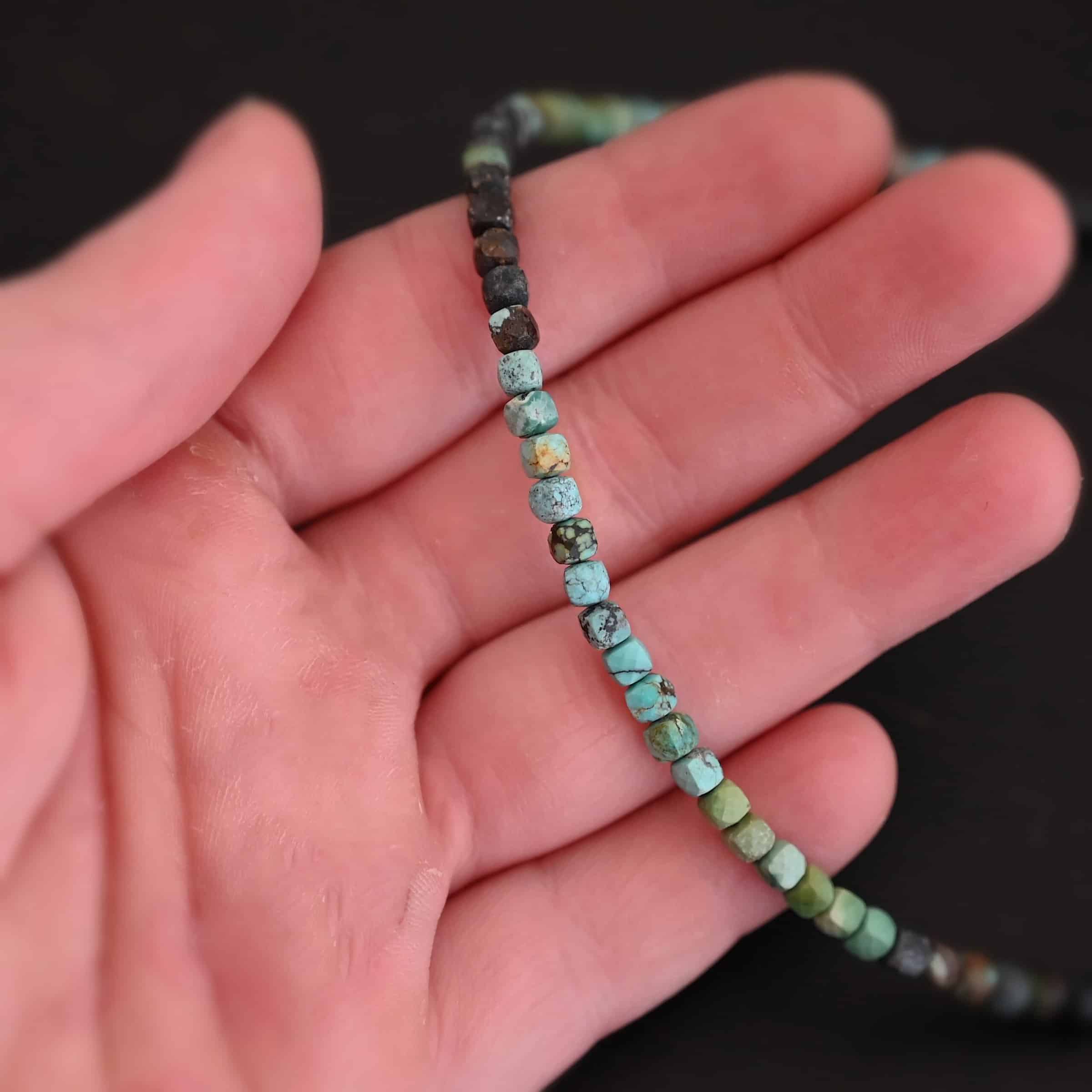 Ada Necklace Close up of the "Ada" Necklace. Necklace is made out of larger sized, square, turquoise beads. The beads are strung on heavy wire and are right next to each other tightly. Beads are a mix of blue, green, black, and brown colors. Necklace is lying against the fingertips of a hand to demonstrate size.