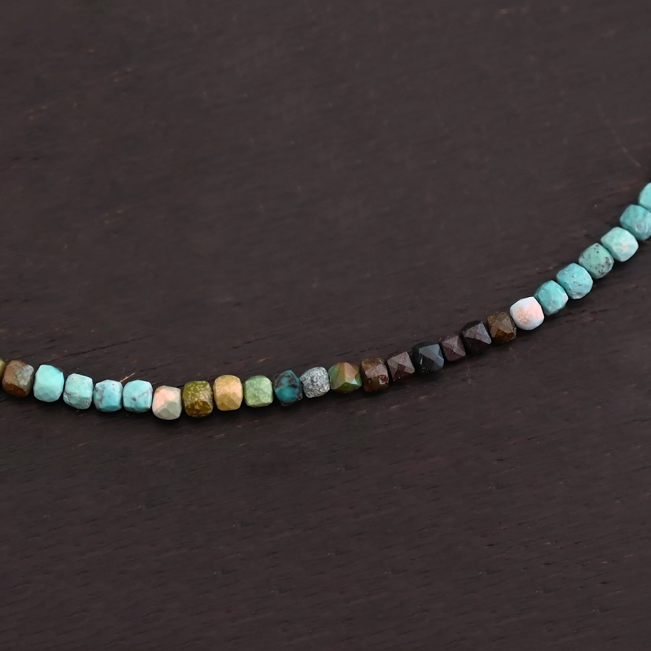 Lenora Necklace Close up of the "Lenora" Necklace. Necklace is made out of smaller sized, square, turquoise beads. The beads are strung on heavy wire and are right next to each other tightly. Beads are a mix of blue, green, black, and brown colors. Necklace is lying against a dark background.