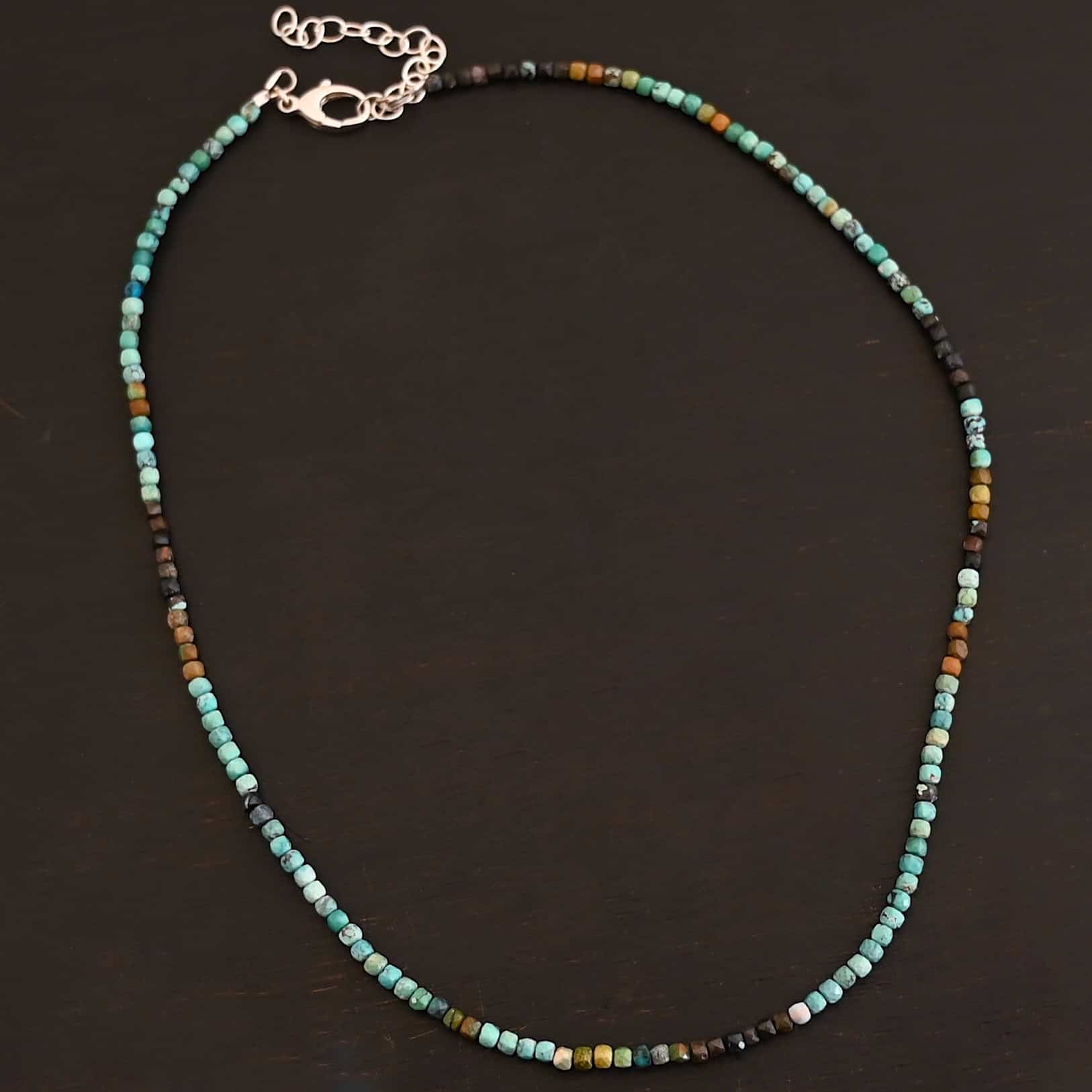 Lenora Necklace Close up of the "Lenora" Necklace. Necklace is made out of smaller sized, square, turquoise beads. The beads are strung on heavy wire and are right next to each other tightly. Beads are a mix of blue, green, black, and brown colors. Necklace has a lobster claw clasp and a two inch sterling silver extension. Necklace is lying against a dark background.