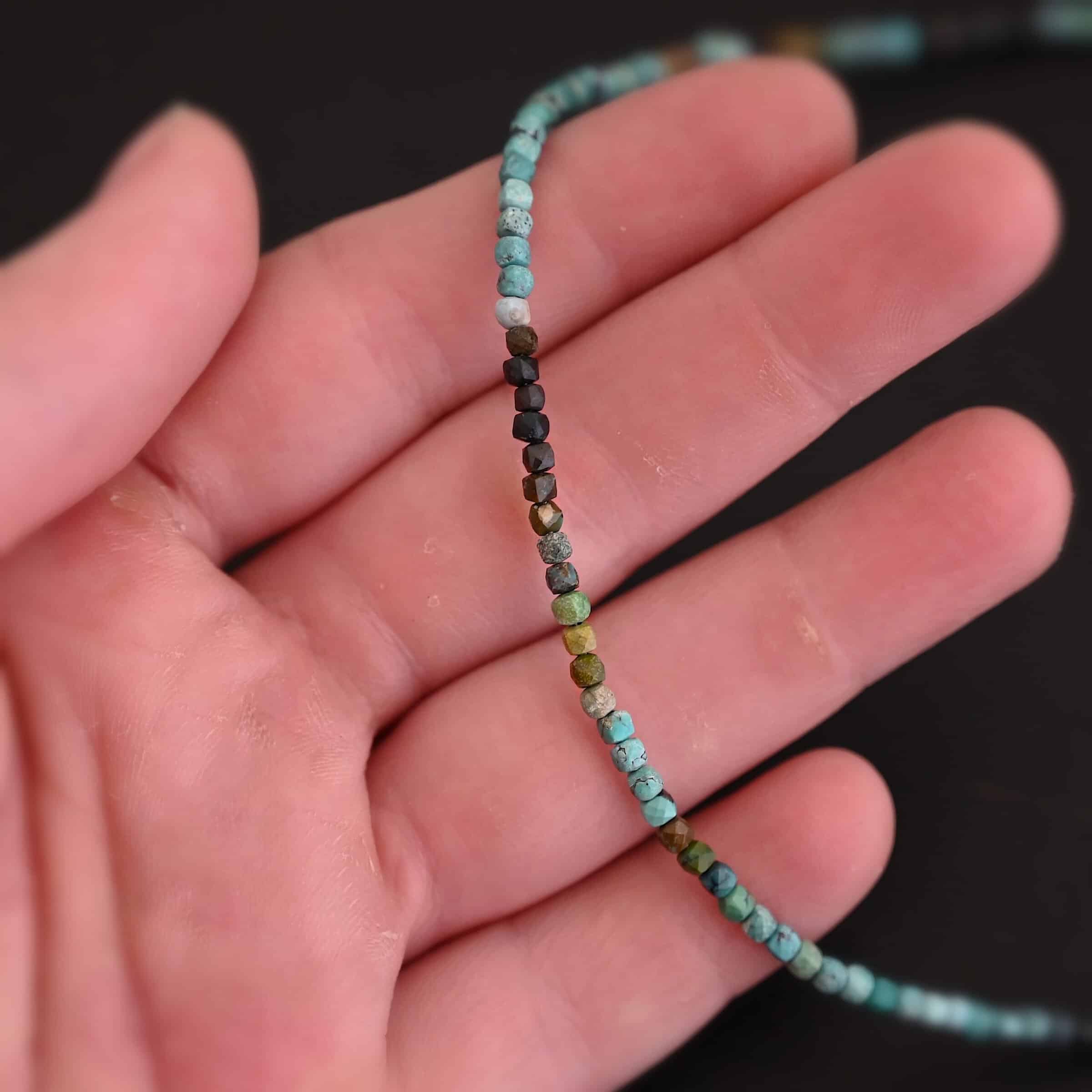Lenora Necklace Close up of the "Lenora" Necklace. Necklace is made out of smaller sized, square, turquoise beads. The beads are strung on heavy wire and are right next to each other tightly. Beads are a mix of blue, green, black, and brown colors. Necklace is lying against the fingertips of a hand to demonstrate size.