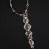 Photo of the "Nightfall" Necklace. Made out of sterling silver, gold, and diamond. There are five diamonds, all in a straight line, that create the pendant of the necklace. Each of the diamonds are different shapes, sizes, and colors. Each of these diamonds are separated by gold balls, which there are six of. The top of the pendant starts with a gold ball, then a large diamond, then a gold ball, and keeps alternating until the final diamond that has a gold ball on the end of it. There is a large loop soldered to the back of the top of the pendant with three gold jump rings attached to it. Going through the jump rings is the silver chain of the necklace. The chain of the necklace has gold loops scattered around it. Necklace is lying against a dark background.