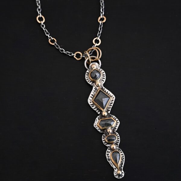 Photo of the "Nightfall" Necklace. Made out of sterling silver, gold, and diamond. There are five diamonds, all in a straight line, that create the pendant of the necklace. Each of the diamonds are different shapes, sizes, and colors. Each of these diamonds are separated by gold balls, which there are six of. The top of the pendant starts with a gold ball, then a large diamond, then a gold ball, and keeps alternating until the final diamond that has a gold ball on the end of it. There is a large loop soldered to the back of the top of the pendant with three gold jump rings attached to it. Going through the jump rings is the silver chain of the necklace. The chain of the necklace has gold loops scattered around it. Necklace is lying against a dark background.