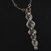 Photo of the "Nightfall" Necklace. Made out of sterling silver, gold, and diamond. There are five diamonds, all in a straight line, that create the pendant of the necklace. Each of the diamonds are different shapes, sizes, and colors. Each of these diamonds are separated by gold balls, which there are six of. The top of the pendant starts with a gold ball, then a large diamond, then a gold ball, and keeps alternating until the final diamond that has a gold ball on the end of it. There is a large loop soldered to the back of the top of the pendant with three gold jump rings attached to it. Going through the jump rings is the silver chain of the necklace. The chain of the necklace has gold loops scattered around it. Necklace is lying against a dark background.