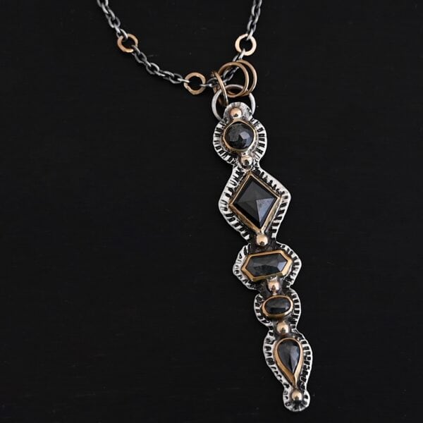 Photo of the "Nightfall" Necklace. Made out of sterling silver, gold, and diamond. There are five diamonds, all in a straight line, that create the pendant of the necklace. Each of the diamonds are different shapes, sizes, and colors. Each of these diamonds are separated by gold balls, which there are six of. The top of the pendant starts with a gold ball, then a large diamond, then a gold ball, and keeps alternating until the final diamond that has a gold ball on the end of it. There is a large loop soldered to the back of the top of the pendant with three gold jump rings attached to it. Going through the jump rings is the silver chain of the necklace. The chain of the necklace has gold loops scattered around it. Necklace is lying against a dark background.