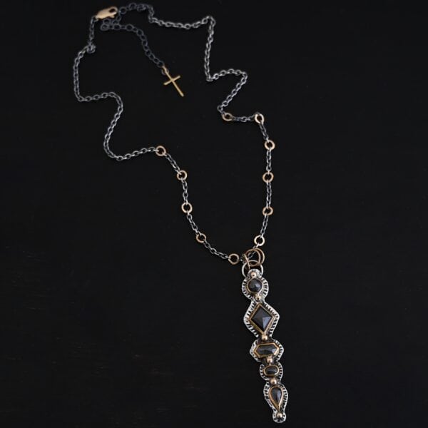 Photo of the "Nightfall" Necklace. Made out of sterling silver, gold, and diamond. There are five diamonds, all in a straight line, that create the pendant of the necklace. Each of the diamonds are different shapes, sizes, and colors. Each of these diamonds are separated by gold balls, which there are six of. The top of the pendant starts with a gold ball, then a large diamond, then a gold ball, and keeps alternating until the final diamond that has a gold ball on the end of it. There is a large loop soldered to the back of the top of the pendant with three gold jump rings attached to it. Going through the jump rings is the silver chain of the necklace. The chain of the necklace has gold loops scattered around it. Necklace is lying against a dark background.