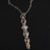 Photo of the "Daybreak" Necklace. Made out of sterling silver, gold, and diamond. There are nine diamonds, all in a straight line, that create the pendant of the necklace. Five of the diamonds are different shapes, sizes, and colors. Each of these diamonds are separated by small, circular diamonds, which there are four of. The top of the pendant starts with a large diamond, then a small one, and keeps alternating until the final diamond is a large one. There is a large loop soldered to the back of the top of the pendant with four gold jump rings attached to it. Going through the jump rings is the silver chain of the necklace. The chain of the necklace has gold loops scattered around it. Necklace is lying against a dark background.