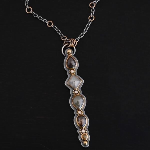 Photo of the "Daybreak" Necklace. Made out of sterling silver, gold, and diamond. There are nine diamonds, all in a straight line, that create the pendant of the necklace. Five of the diamonds are different shapes, sizes, and colors. Each of these diamonds are separated by small, circular diamonds, which there are four of. The top of the pendant starts with a large diamond, then a small one, and keeps alternating until the final diamond is a large one. There is a large loop soldered to the back of the top of the pendant with four gold jump rings attached to it. Going through the jump rings is the silver chain of the necklace. The chain of the necklace has gold loops scattered around it. Necklace is lying against a dark background.