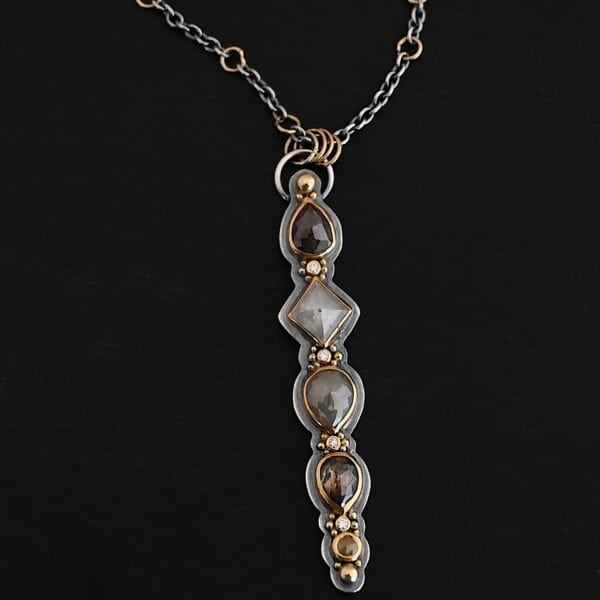 Photo of the "Daybreak" Necklace. Made out of sterling silver, gold, and diamond. There are nine diamonds, all in a straight line, that create the pendant of the necklace. Five of the diamonds are different shapes, sizes, and colors. Each of these diamonds are separated by small, circular diamonds, which there are four of. The top of the pendant starts with a large diamond, then a small one, and keeps alternating until the final diamond is a large one. There is a large loop soldered to the back of the top of the pendant with four gold jump rings attached to it. Going through the jump rings is the silver chain of the necklace. The chain of the necklace has gold loops scattered around it. Necklace is lying against a dark background.