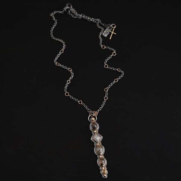 Photo of the "Daybreak" Necklace. Made out of sterling silver, gold, and diamond. There are nine diamonds, all in a straight line, that create the pendant of the necklace. Five of the diamonds are different shapes, sizes, and colors. Each of these diamonds are separated by small, circular diamonds, which there are four of. The top of the pendant starts with a large diamond, then a small one, and keeps alternating until the final diamond is a large one. There is a large loop soldered to the back of the top of the pendant with four gold jump rings attached to it. Going through the jump rings is the silver chain of the necklace. The chain of the necklace has gold loops scattered around it. Necklace is lying against a dark background.