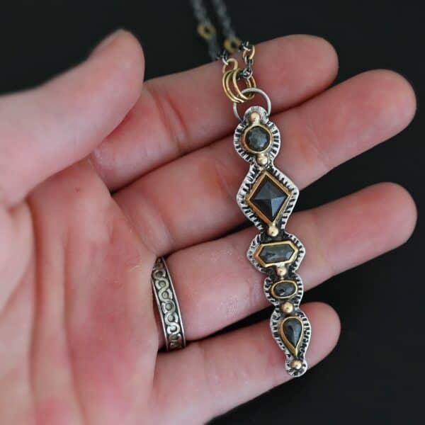Photo of the "Nightfall" Necklace. Made out of sterling silver, gold, and diamond. There are five diamonds, all in a straight line, that create the pendant of the necklace. Each of the diamonds are different shapes, sizes, and colors. Each of these diamonds are separated by gold balls, which there are six of. The top of the pendant starts with a gold ball, then a large diamond, then a gold ball, and keeps alternating until the final diamond that has a gold ball on the end of it. There is a large loop soldered to the back of the top of the pendant with three gold jump rings attached to it. Going through the jump rings is the silver chain of the necklace. The chain of the necklace has gold loops scattered around it. Necklace is lying against the fingertips of a hand to demonstrate size.