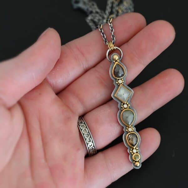 Photo of the "Daybreak" Necklace. Made out of sterling silver, gold, and diamond. There are nine diamonds, all in a straight line, that create the pendant of the necklace. Five of the diamonds are different shapes, sizes, and colors. Each of these diamonds are separated by small, circular diamonds, which there are four of. The top of the pendant starts with a large diamond, then a small one, and keeps alternating until the final diamond is a large one. There is a large loop soldered to the back of the top of the pendant with four gold jump rings attached to it. Going through the jump rings is the silver chain of the necklace. The chain of the necklace has gold loops scattered around it. Necklace is lying against the fingertips of a hand to demonstrate size.