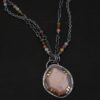 Photo of the "Peach Tree" Necklace. Made out of sterling silver, gold, peach moonstone, and tourmaline. There is a rose cut, octagon shaped peach moonstone in the center with eight gold prongs holding the stone in. The prongs are four groups of two in each corner of the ring. The silver around the stone has hammered marks in groups scattered around it as well. There is a loop on the back of the pendant with three silver jump rings going through it. Going through the jump rings is the chain of the necklace. The chain of the necklace is two separate chains that meet at the back to make one chain for the necklace. Each chain has multiple tourmaline beads on it. Necklace is lying against a dark background.