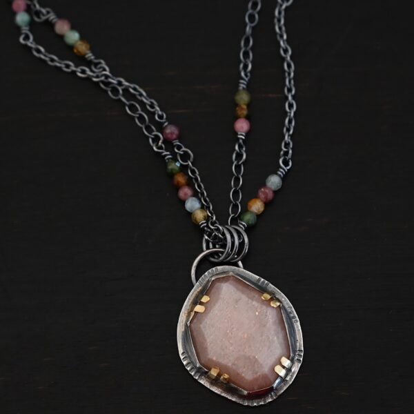 Photo of the "Peach Tree" Necklace. Made out of sterling silver, gold, peach moonstone, and tourmaline. There is a rose cut, octagon shaped peach moonstone in the center with eight gold prongs holding the stone in. The prongs are four groups of two in each corner of the ring. The silver around the stone has hammered marks in groups scattered around it as well. There is a loop on the back of the pendant with three silver jump rings going through it. Going through the jump rings is the chain of the necklace. The chain of the necklace is two separate chains that meet at the back to make one chain for the necklace. Each chain has multiple tourmaline beads on it. Necklace is lying against a dark background.
