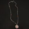 Photo of the "Peach Tree" Necklace. Made out of sterling silver, gold, peach moonstone, and tourmaline. There is a rose cut, octagon shaped peach moonstone in the center with eight gold prongs holding the stone in. The prongs are four groups of two in each corner of the ring. The silver around the stone has hammered marks in groups scattered around it as well. There is a loop on the back of the pendant with three silver jump rings going through it. Going through the jump rings is the chain of the necklace. The chain of the necklace is two separate chains that meet at the back to make one chain for the necklace. Each chain has multiple tourmaline beads on it. Necklace is lying against a dark background.