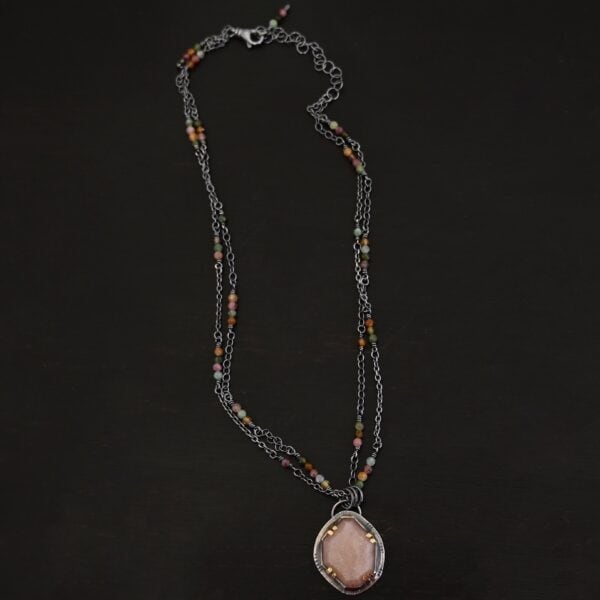 Photo of the "Peach Tree" Necklace. Made out of sterling silver, gold, peach moonstone, and tourmaline. There is a rose cut, octagon shaped peach moonstone in the center with eight gold prongs holding the stone in. The prongs are four groups of two in each corner of the ring. The silver around the stone has hammered marks in groups scattered around it as well. There is a loop on the back of the pendant with three silver jump rings going through it. Going through the jump rings is the chain of the necklace. The chain of the necklace is two separate chains that meet at the back to make one chain for the necklace. Each chain has multiple tourmaline beads on it. Necklace is lying against a dark background.