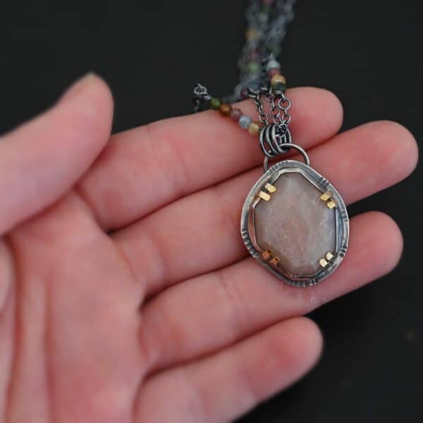 Photo of the "Peach Tree" Necklace. Made out of sterling silver, gold, peach moonstone, and tourmaline. There is a rose cut, octagon shaped peach moonstone in the center with eight gold prongs holding the stone in. The prongs are four groups of two in each corner of the ring. The silver around the stone has hammered marks in groups scattered around it as well. There is a loop on the back of the pendant with three silver jump rings going through it. Going through the jump rings is the chain of the necklace. The chain of the necklace is two separate chains that meet at the back to make one chain for the necklace. Each chain has multiple tourmaline beads on it. Necklace is lying against the fingertips of a hand to demonstrate size.