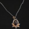 Photo of the "Golden Fields" Necklace. Made out of sterling silver, gold, sapphire, and diamonds. The center stone is an oval shaped, deep brown sapphire that is set in gold. There is a gold ball on the left and right side of this sapphire. There are also four gold leaves, two on each side, beneath the gold balls. The ends of each leaf meet, and where they meet in the center is a gold ball. There are three diamonds, two small and one big, beneath the sapphire, that the gold leaves surround. The two small diamonds are on the side with the big one in the middle. There are three more gold balls that surround each side of the big diamond. There is a loop on the back of the necklace, with three gold jump rings that are attached to it. Going through the jump rings is the chain of the necklace. Necklace is lying against a dark background.