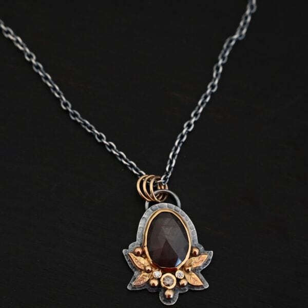 Photo of the "Golden Fields" Necklace. Made out of sterling silver, gold, sapphire, and diamonds. The center stone is an oval shaped, deep brown sapphire that is set in gold. There is a gold ball on the left and right side of this sapphire. There are also four gold leaves, two on each side, beneath the gold balls. The ends of each leaf meet, and where they meet in the center is a gold ball. There are three diamonds, two small and one big, beneath the sapphire, that the gold leaves surround. The two small diamonds are on the side with the big one in the middle. There are three more gold balls that surround each side of the big diamond. There is a loop on the back of the necklace, with three gold jump rings that are attached to it. Going through the jump rings is the chain of the necklace. Necklace is lying against a dark background.