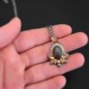 Photo of the "Golden Fields" Necklace. Made out of sterling silver, gold, sapphire, and diamonds. The center stone is an oval shaped, deep brown sapphire that is set in gold. There is a gold ball on the left and right side of this sapphire. There are also four gold leaves, two on each side, beneath the gold balls. The ends of each leaf meet, and where they meet in the center is a gold ball. There are three diamonds, two small and one big, beneath the sapphire, that the gold leaves surround. The two small diamonds are on the side with the big one in the middle. There are three more gold balls that surround each side of the big diamond. There is a loop on the back of the necklace, with three gold jump rings that are attached to it. Going through the jump rings is the chain of the necklace. Necklace is lying against the fingertips of a hand to demonstrate size.