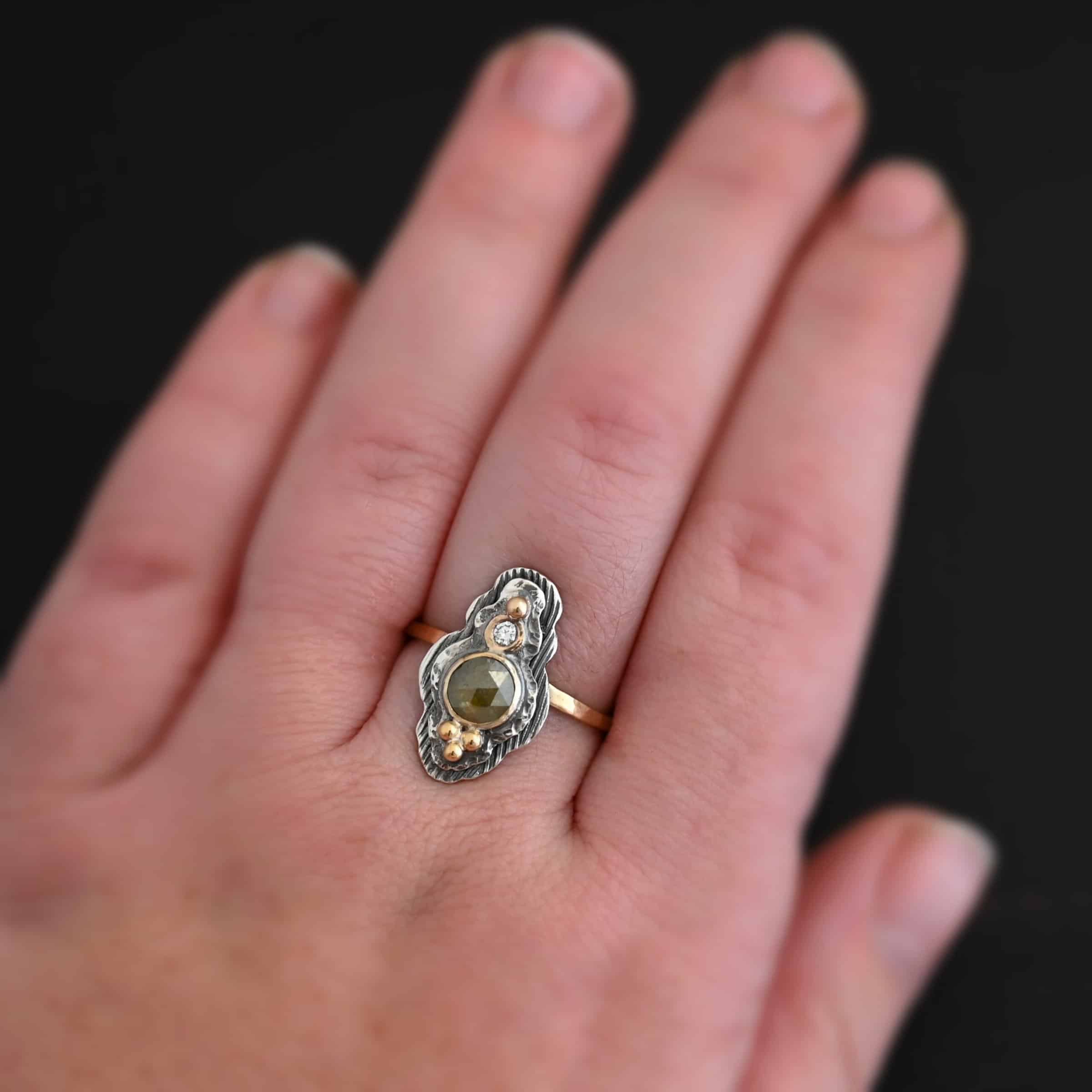 Garden Of Eden Ring Photo of the "Garden Of Eden" Ring. Made out of sterling silver and diamonds. There is a ball of gold on the top and a small, clear, circular diamond beneath it. Then, a larger, darker green, circular diamond, and three balls of gold beneath that in a triangle shape. There are two layers of silver, both with hammered lines in them. The band is thin gold that is hammered. Ring is worn by a model.