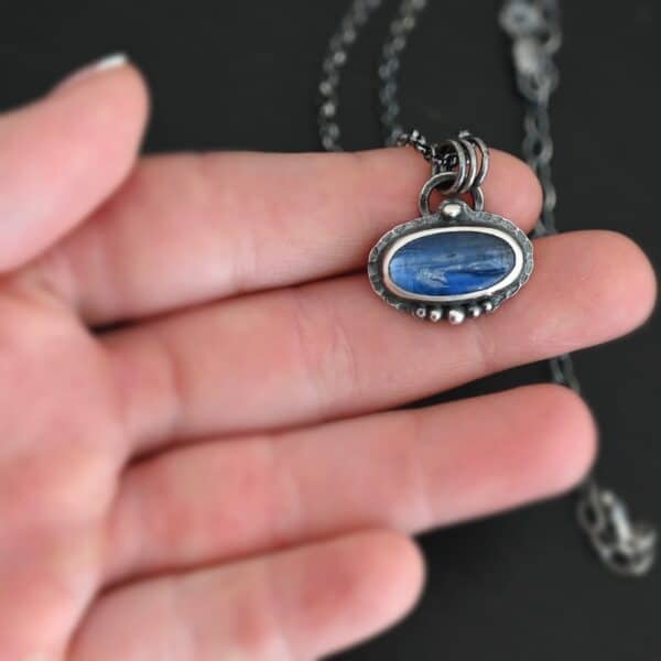 Photo of the "Caelum" Necklace. Made out of sterling silver and kyanite. There is an oval shaped deep blue kyanite set horizontally in the center. There is a ball of silver on the top of it in the center. There are five more balls of silver beneath it, that go in a straight line across the bottom. The rest of the silver around it is hammered. There is a loop soldered to the back with three jump rings attached to it. The chain for the necklace goes through the jump rings. Necklace is lying on the fingertips of a hand.